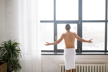Rear view of guy with towel on hips looking through window after having bath