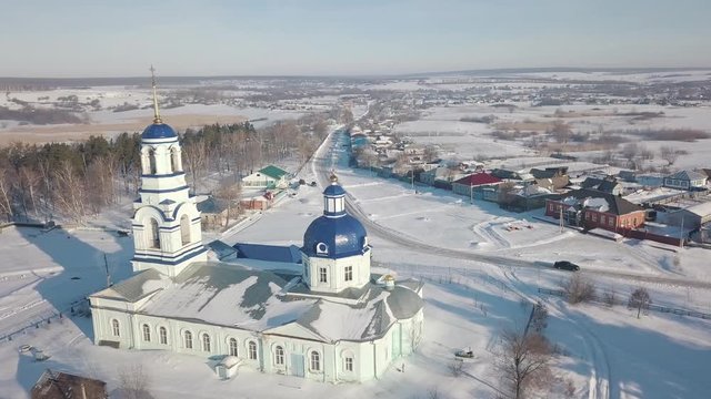 Aerial view of the white Russian orthodox church with blue domes and golden crosses located near the small village in sunny winter day. Shot. Religion and faith