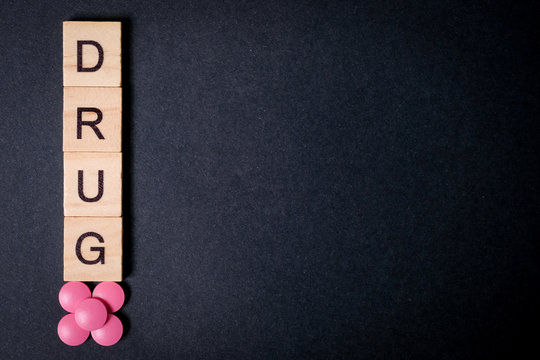 The word drug is on wooden squares next to pink tablets on a black background. Copy space. Top view.
