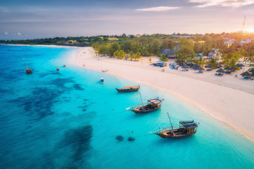 Aerial view of the fishing boats on tropical sea coast with white sandy beach at sunset. Summer holiday on Indian Ocean, Zanzibar. Landscape with boat, palm trees, transparent blue water. Top view - obrazy, fototapety, plakaty