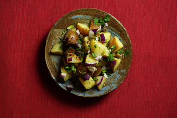 Fototapeta na wymiar Potato salad with Dijon dressing along with parsley, dill, red onion and capers. Textured red napkin. Top view, horizontal arrangement.