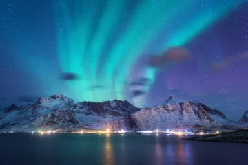 Zelfklevend Fotobehang Aurora borealis over de zee, besneeuwde bergen en stadslichten & 39 s nachts. Noorderlicht op de Lofoten-eilanden, Noorwegen. Paarse sterrenhemel met poollicht. Winterlandschap met aurora weerspiegeld in water © den-belitsky