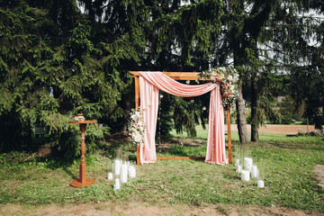 View over decorated wedding arch with coral draped cloth, flowers and big candles on the ground in...