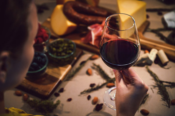 Holding a glass of red wine and in the background a table with gourmet ingredients, variety of cheeses, cold meats, grapes, raspberry, blackberry and artisanal bread on a rustic background