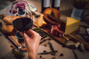 Holding a glass of red wine and in the background a table with gourmet ingredients, variety of cheeses, cold meats, grapes, raspberry, blackberry and artisanal bread on a rustic background