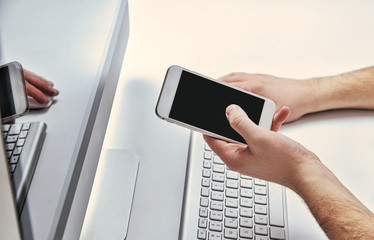 Man holds phone in hand in front of computer at home, office. Concept of using technology in business, home. Smartphone in hand in front of the monitor. Checking information on the internet, payment.