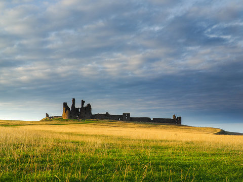 Dramatic View of the Ruins of Dunstanburgh Castle on a Hilltop in Northumberland, England, Surrounded by Golden and Green Grass Fields under a Cloudy Sky at Sunrise or Sunset