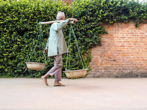 A Traditional Nepali Porter Walking Barefoot and Carrying Baskets Balanced on a Yoke Against a Green and Brick Wall Background