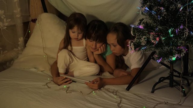 concept of family Christmas. children on Christmas evening play on tablet, in children's room on couch, in tent with garlands. sisters play in a room decorated with a multi-colored New Year tree