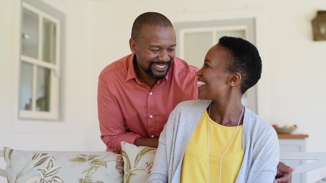 Happy mature black couple bonding to each other and smiling while sitting on couch. Portrait of smiling black man embrace his wife from behind and looking at camera. Husband and woman laughing.