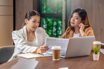 Two asian businesswomen working with the partner business with paper graph document and technology laptop in modern meeting room, office or working space, coffee break, partner and colleague concept