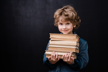 Successful schoolboy holding stack of books while standing in front of camera