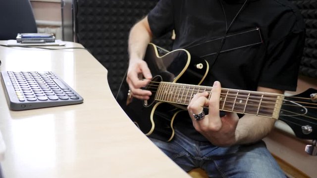 Guitarist Playing Acoustic Guitar at Home Recording Studio. Man dressed in black T-shirt plays stringed musical instrument. Desk with computer keyboard. Soundproof walls background