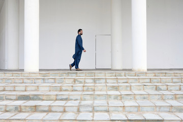 Middle east man with beard wearing blue suit walking on the white building with stair steps concerte poles and white wall background, Bahrian