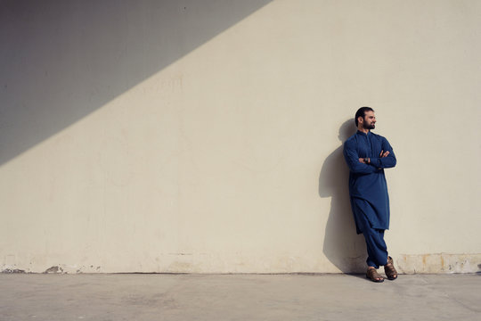 Young Man With Beard Wearing Blue Long Traditional Cloth Standing At Wall Wtih Sunlight And Shadow Background, Isolated.