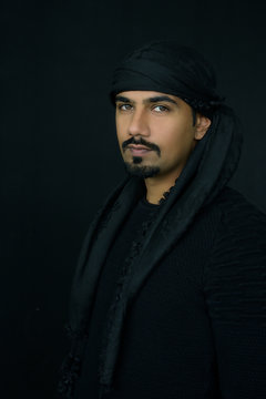 Handsome middle east man with beard wearing long black dress with turban standing in dark room background, Bahrain.