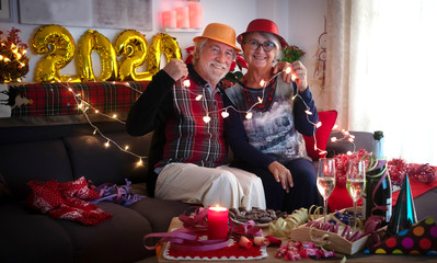 Two cheerful senior people with a shimmering hat sitting on the sofa celebrating the new year. Toasting with a sparkling wine. Lights and decorations around them