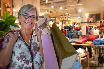 Front view of a senior woman smiling and satisfied with purchases in a mall. Gray hair and eyeglasses. Loaded with shopping bags