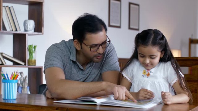 Indian father wearing spectacles reading story book with daughter. HD Stock Video of happy father-daughter doing fun activities in the living room. Parents spending quality time with children - Rea...