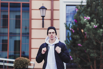 Young beautiful fashionable guy posing in street and wearing backpack, jacket, glasses, urban style