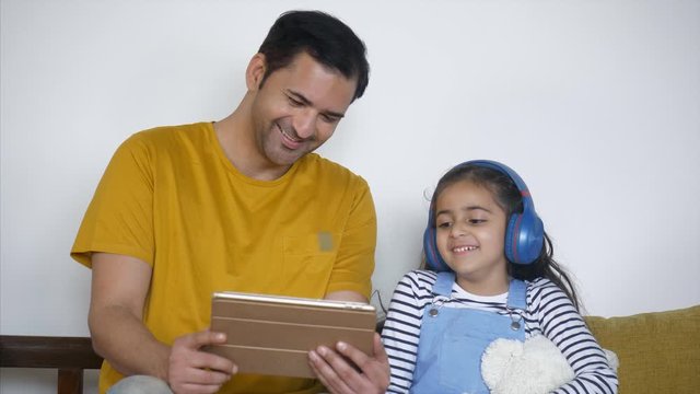 Cute little girl happily watching cartoons on a digital tablet with her father - weekend fun. Indian dad showing funny videos to his beautiful daughter on an iPad while sitting on a couch - technol...