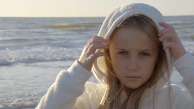 Girl blonde 8 year old cover her head with a white hood, background of the sea