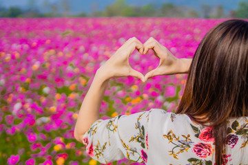 Obraz na płótnie Canvas The girl raised her hand to display a meaningful heart-shaped symbol. Tell her that I love you on the blurred background of the flower garden. Showing symbols to tell love Lover on valentine's day