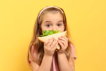 Tuinposter Cute little girl eating tasty sandwich on yellow background © New Africa
