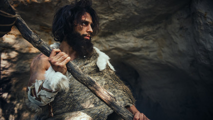 Fototapeta na wymiar Primeval Caveman Wearing Animal Skin Holds Stone Hammer Comes out of Cave and Looks Around Prehistoric Landscape, Ready to Hunt Animal Prey. Neanderthal Going to Hunt in a Jungle. Low Angle Shot
