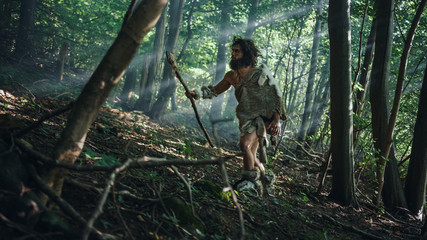 Naklejka na ściany i meble Primeval Caveman Wearing Animal Skin Holds Stone Tipped Spear Looks Around, Explores Prehistoric Forest in a Hunt for Animal Prey. Neanderthal Going Hunting in the Jungle
