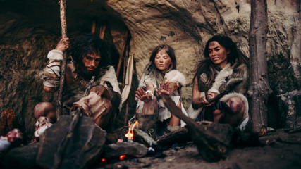 Obraz na płótnie Canvas Tribe of Prehistoric PrimitiveHunter-Gatherers Wearing Animal Skins Live in a Cave at Night. Neanderthal or Homo Sapiens Family Trying to Get Warm at the Bonfire, Holding Hands over Fire, Cooking Food