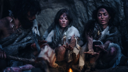 Tribe of Prehistoric Hunter-Gatherers Wearing Animal Skins Live in Cave at Night. Neanderthal Family Trying to Get Warm at the Bonfire, Holding Hands over Fire, Cooking Food. Shot with a Warm Filter.