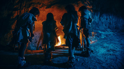 Tribe of Prehistoric Hunter-Gatherers Wearing Animal Skins Live in a Cave at Night. Neanderthal or Homo Sapiens Family Trying to Get Warm at the Bonfire, Holding Hands over Fire. Back View