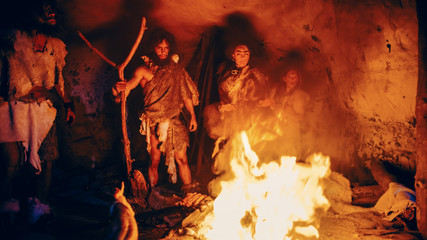 Tribe of Prehistoric Hunter-Gatherers Wearing Animal Skins Stand Around Bonfire Outside of Cave at...