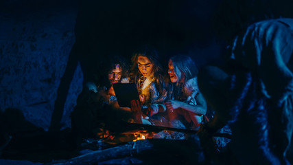 Tribe of Prehistoric, Primitive Hunter-Gatherers Wearing Animal Skins Use Digital Tablet Computer in a Cave at Night. Neanderthal or Homo Sapiens Family Browsing Internet, Watching Videos, Streaming 