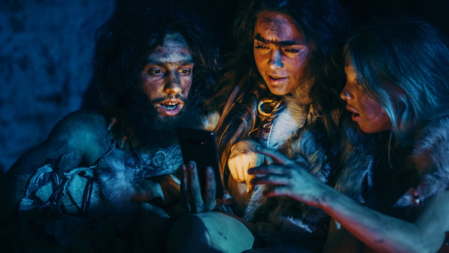 Tribe Of Prehistoric, Primitive Hunter-Gatherers Wearing Animal Skins Use Smartphone In A Cave At Night. Neanderthal / Homo Sapiens Family Browsing Internet On Mobile Phone