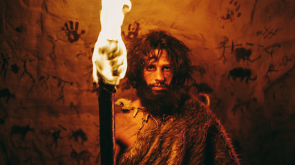Portrait of Primeval Caveman Wearing Animal Skin Standing in His Cave At Night, Holding Torch with...