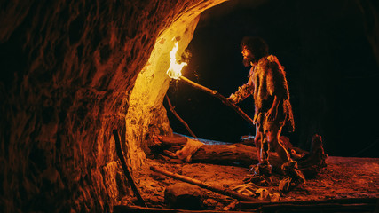 Primeval Caveman Wearing Animal Skin Exploring Cave At Night, Holding Torch with Fire Looking at...