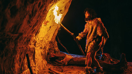 Primeval Caveman Wearing Animal Skin Exploring Cave At Night, Holding Torch with Fire Looking at...