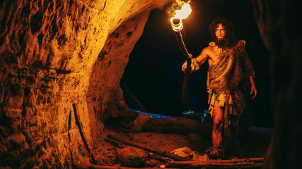 Primeval Caveman Wearing Animal Skin Exploring Cave At Night, Holding Torch with Fire Looking at Drawings on the Walls at Night. Neanderthal Searching Safe Place to Spend the Night - obrazy, fototapety, plakaty