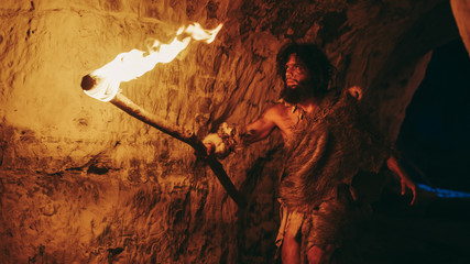 Primeval Caveman Wearing Animal Skin Exploring Cave At Night, Holding Torch with Fire Looking at...