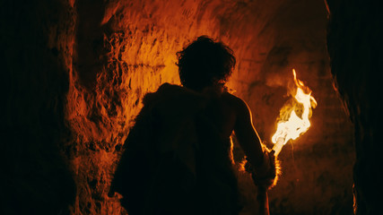 Primeval Caveman Wearing Animal Skin Exploring Cave At Night Holding Torch with Fire Looking at...