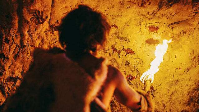 Primeval Caveman Wearing Animal Skin Standing in His Cave At Night, Holding Torch with Fire Looking at Drawings on the Walls at Night. Cave Art with Petroglyphs, Rock Paintings. Back View