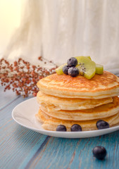 Stack of pancakes with topping, kiwi and blueberry.placed in a white plate on a blue wooden table.Eat with milk tea in the glass.