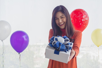 Beautiful woman with long Asian hair wearing a red sweater, smiling happy in New Year's Eve and Christmas.She holds a gift to show off wrapped in yellow paper.Background of balloons of various colors.
