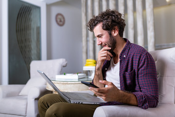 Successful entrepreneur smiling in satisfaction as he checks information on his laptop computer while working in a home office. Young man relaxing on the sofa with a laptop and mobile phone in hands