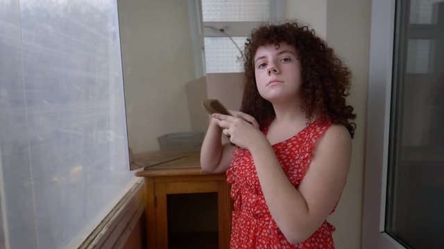 cute freckled plump teenage girl combing her curly brunette hair in front of a mirror in natural light wearing a red dress, slow motion 4k 