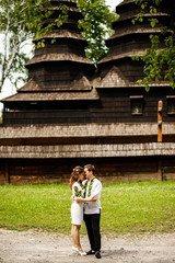 Lovely newlyweds in embroidered costumes stand near an old wooden church