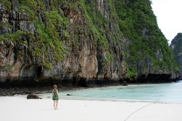 A girl with red hair in a green dress stands on the seashore and looks into the distance. A sea of ​​amazing azure color. The girl stands next to a huge rocky back to the viewer.