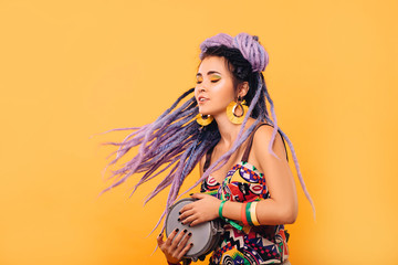 Hipster woman with violet dreadlocks and colored clothes playing on ethical mini drum on yellow...